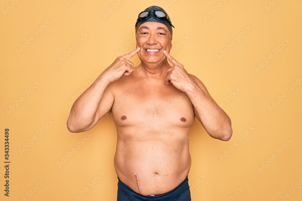 Middle age senior grey-haired swimmer man wearing swimsuit, cap and goggles Smiling with open mouth, fingers pointing and forcing cheerful smile