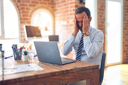 Middle age handsome businessman wearing tie sitting using laptop at the office suffering from headache desperate and stressed because pain and migraine. Hands on head.