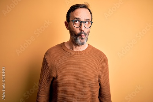 Middle age hoary man wearing brown sweater and glasses over isolated yellow background making fish face with lips, crazy and comical gesture. Funny expression.