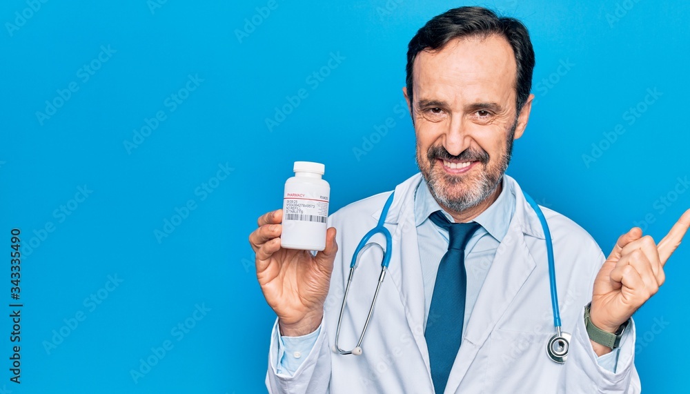 Middle age handsome doctor man wearing stethoscope holding bottle of pills smiling happy pointing with hand and finger to the side Middle age handsome doctor man wearing stethoscope holding bottle of pills smiling happy pointing with hand and finger to the side