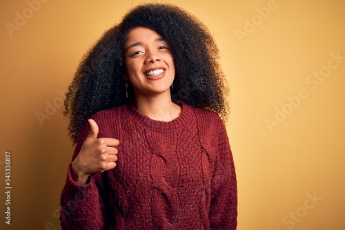 Young beautiful african american woman with afro hair standing over yellow isolated background doing happy thumbs up gesture with hand. Approving expression looking at the camera showing success. © Krakenimages.com