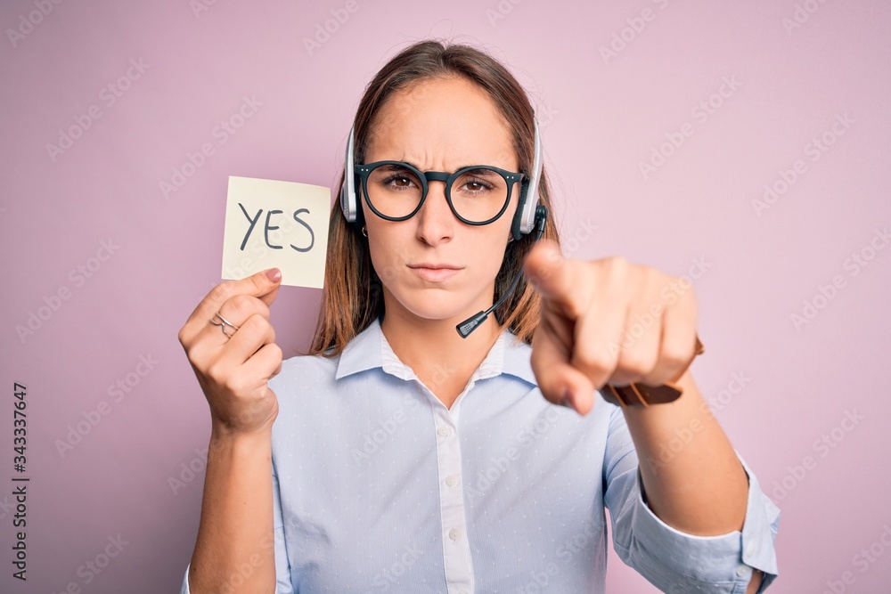 Beautiful call center agent woman working using headset holding reminder with yes message pointing with finger to the camera and to you, hand sign, positive and confident gesture from the front Beautiful call center agent woman working using headset holding reminder with yes message pointing with finger to the camera and to you, hand sign, positive and confident gesture from the front