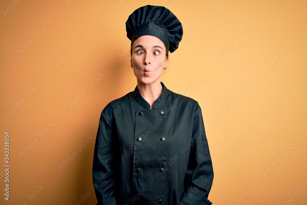 Young beautiful chef woman wearing cooker uniform and hat standing over yellow background making fish face with lips, crazy and comical gesture. Funny expression. Young beautiful chef woman wearing cooker uniform and hat standing over yellow background making fish face with lips, crazy and comical gesture. Funny expression.