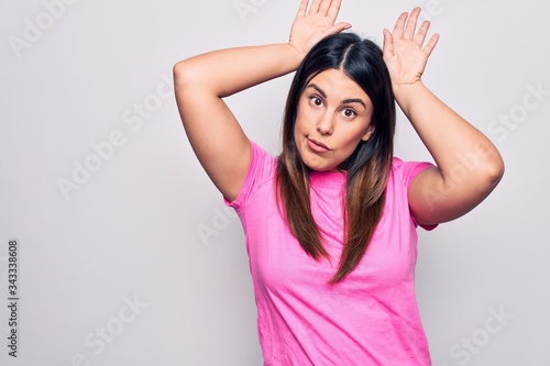 Young beautiful brunette woman wearing casual pink t-shirt standing over white background Doing bunny ears gesture with hands palms looking cynical and skeptical. Easter rabbit concept.