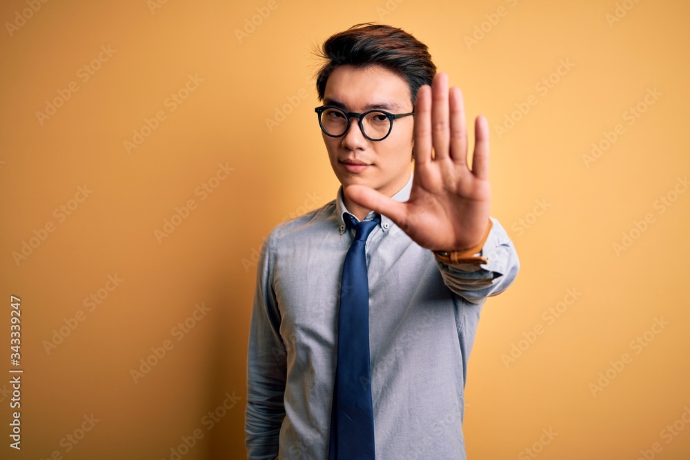 Young handsome chinese businessman wearing glasses and tie over yellow background doing stop sing with palm of the hand. Warning expression with negative and serious gesture on the face. Young handsome chinese businessman wearing glasses and tie over yellow background doing stop sing with palm of the hand. Warning expression with negative and serious gesture on the face.