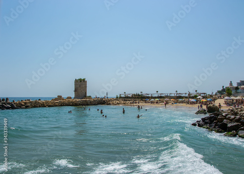 Seascape, view of a beach full with people, old tower of a castle and rocks and tetrapods, blue water and sky, summer season, nature