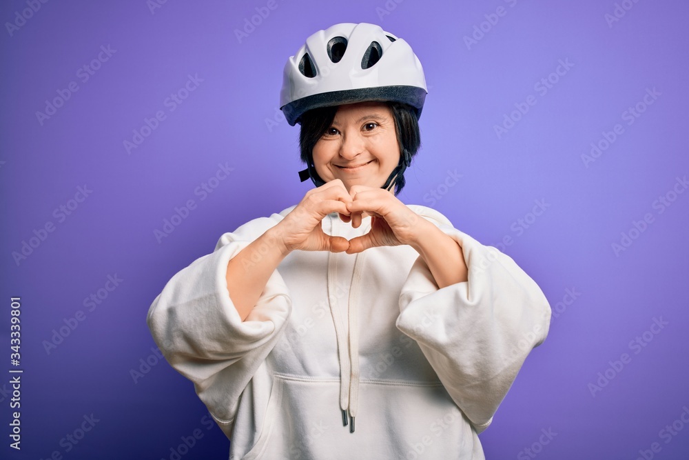 Young down syndrome cyclist woman wearing security bike helmet over purple background smiling in love showing heart symbol and shape with hands. Romantic concept. Young down syndrome cyclist woman wearing security bike helmet over purple background smiling in love showing heart symbol and shape with hands. Romantic concept.