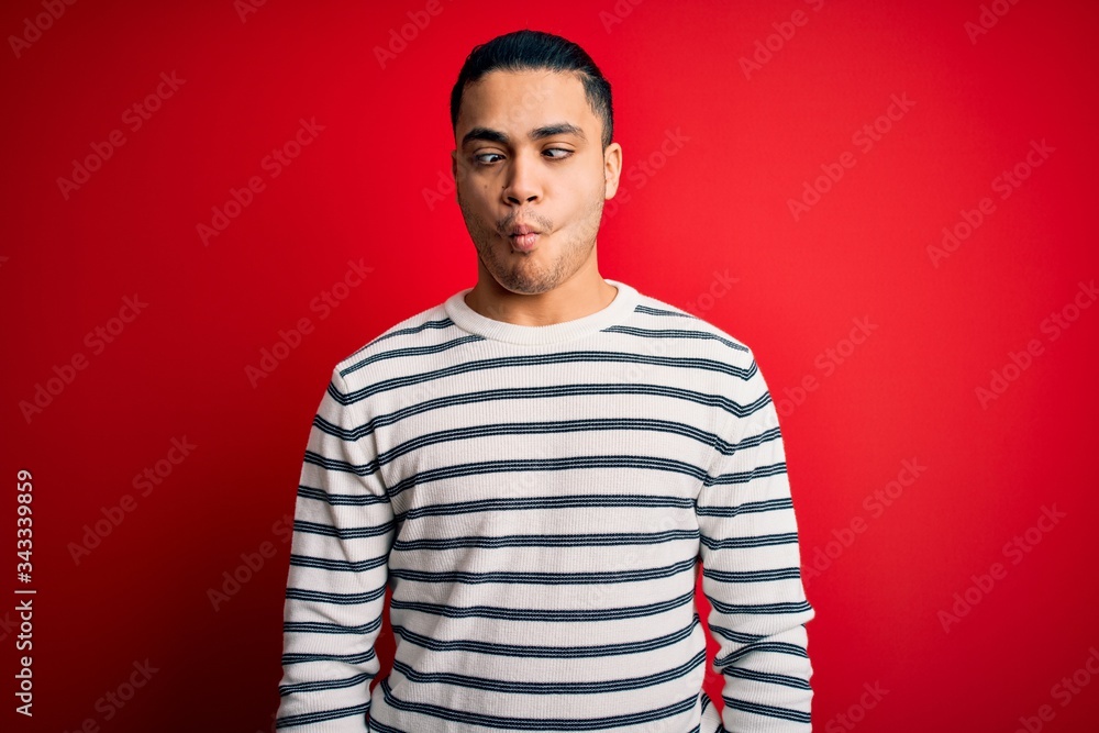 Young brazilian man wearing casual striped t-shirt standing over isolated red background making fish face with lips, crazy and comical gesture. Funny expression. Young brazilian man wearing casual striped t-shirt standing over isolated red background making fish face with lips, crazy and comical gesture. Funny expression.