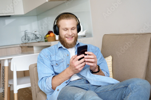 Happy young guy wearing wireless headphones listening to music, audio book or learning language on smartphone in app sit on sofa. Relaxed man enjoying mobile player podcast lounge on couch at home.