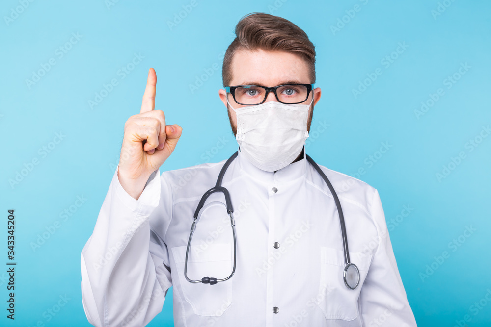 Portrait of doctor wearing a mask and white uniform pointing up with a finger on blue background. Attention, coronavirus outbreak, pandemic and covid-19.
