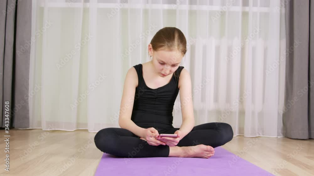 Fitness girl chatting in mobile phone on gym carpet yoga mat at home. Sport girl teenager looking smartphone sitting on fitness carpet while break on home training Fitness girl chatting in mobile phone on gym carpet yoga mat at home. Sport girl teenager looking smartphone sitting on fitness carpet while break on home training