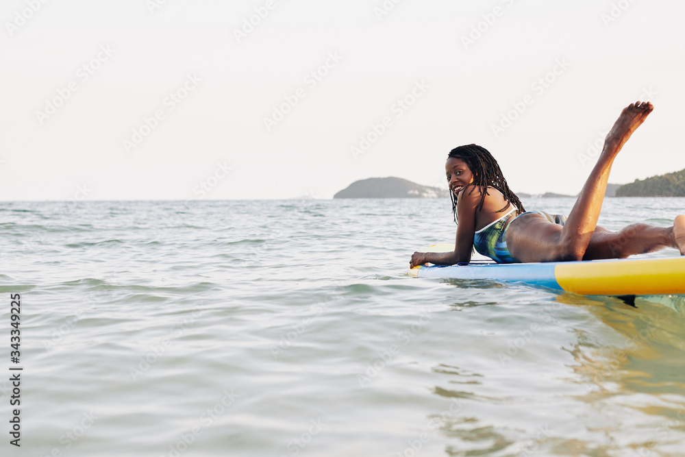 Happy attractive young Black woman lying on sup surfing board and looking at camera