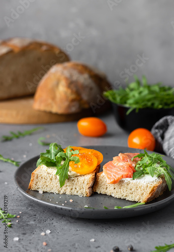 Open sandwiches with wheat and rye bread, tomatoes, smoked salmon and arugula, grey concrete background. Selective focus. 