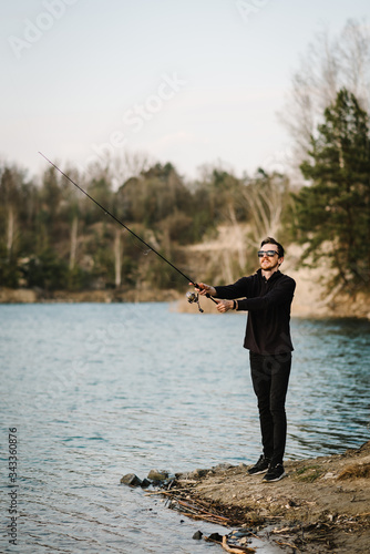 Fisherman with rod, spinning reel on river bank. Sunrise. Man catching fish, pulling rod while fishing from lake or pond. Fishing for pike, perch, carp on beach lake or pond. Background wild nature.