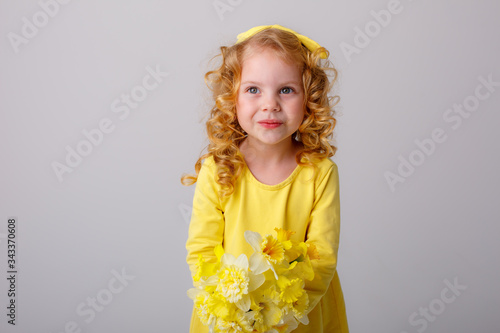 a small curly haired girl in a yellow dress holding a bouquet of spring flowers on a white background smiles