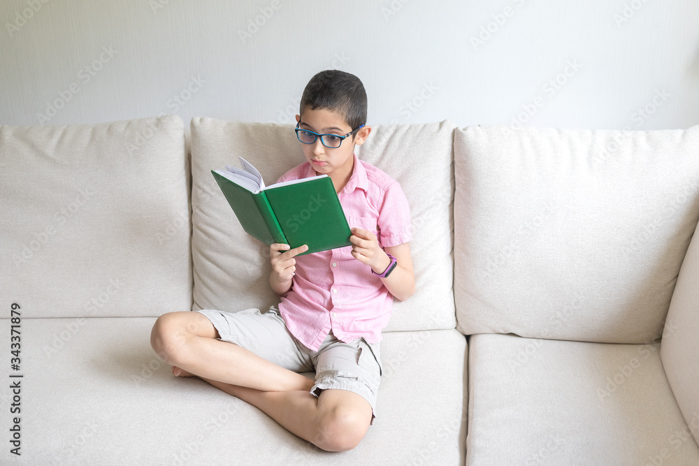 Boy in pink t-shirt sits at home on cozy sofa, reads book. Light background. Quarantine. Holidays. Boy in pink t-shirt sits at home on cozy sofa, reads book. Light background. Quarantine. Holidays.