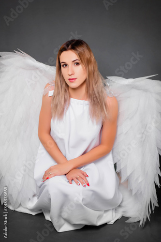 Young woman in a white dress with white wings behind her back. Girl in the image of an angel isolated on a gray background.