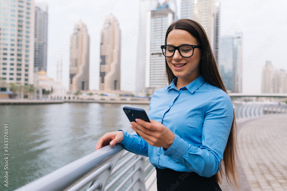 Fototapeta premium Young pretty business woman texting on a mobile phone outdoors on skyscrapers background