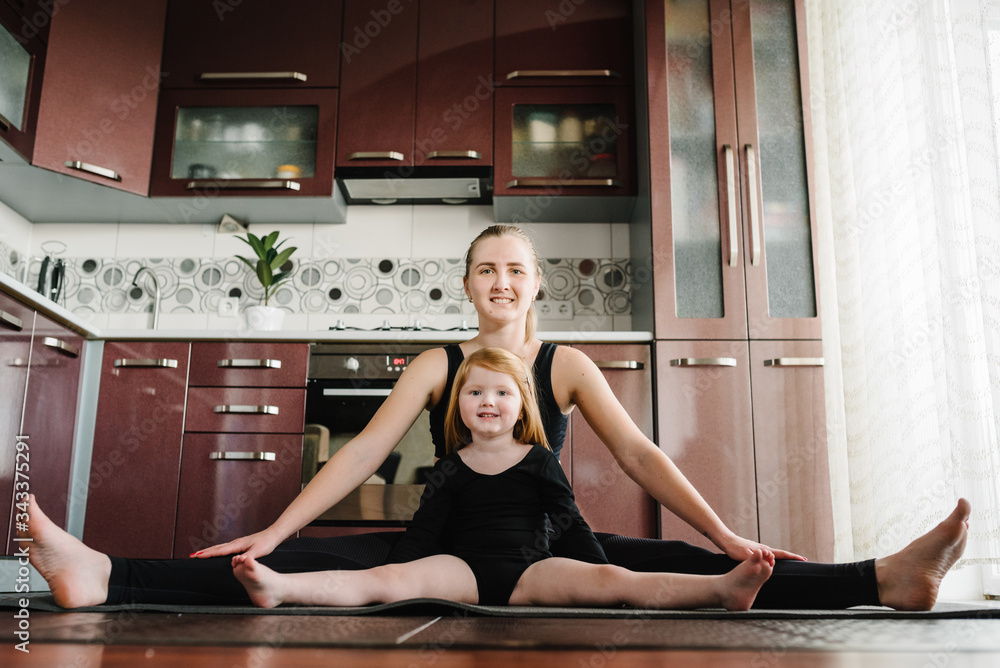 Mother and daughter in tracksuits are training on mat at home. Happy family playing sports in the kitchen. Sports mom with child doing morning work-out, do the exercises together. Stay at home. Mother and daughter in tracksuits are training on mat at home. Happy family playing sports in the kitchen. Sports mom with child doing morning work-out, do the exercises together. Stay at home.