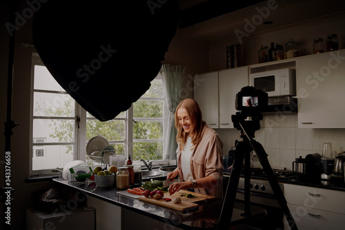 Female food vlogger recording video on dslr camera while chopping vegetables in kitchen in isolation at home in quarantine