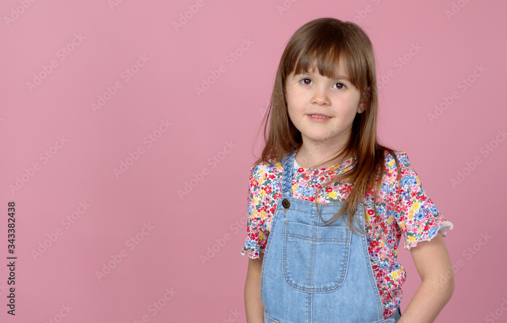 Sweet little girl 6-7 years old posing in dungarees jeans and flower pattern blouse on pink Sweet little girl 6-7 years old posing in dungarees jeans and flower pattern blouse on pink