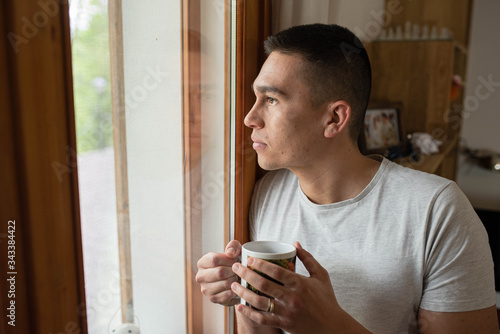Tableau sur toile Casual young man at home holding a cup of coffee and looking out the window with his image reflected on the glass