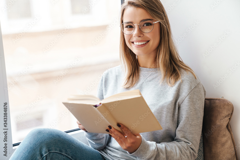 Photo of smiling woman reading book while sitting near window Photo of smiling woman reading book while sitting near window