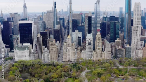 Aerial, tracking, drone shot over the central park, tilting towards skyscrapers and office buildings, cloudy, spring day, in midtown Manhattan, NYC, USA