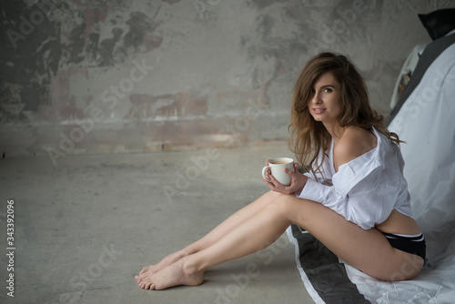 Young beautiful woman with slim long legs in the morning sits on the concrete floor with a cup of coffee. Soft selective focus.