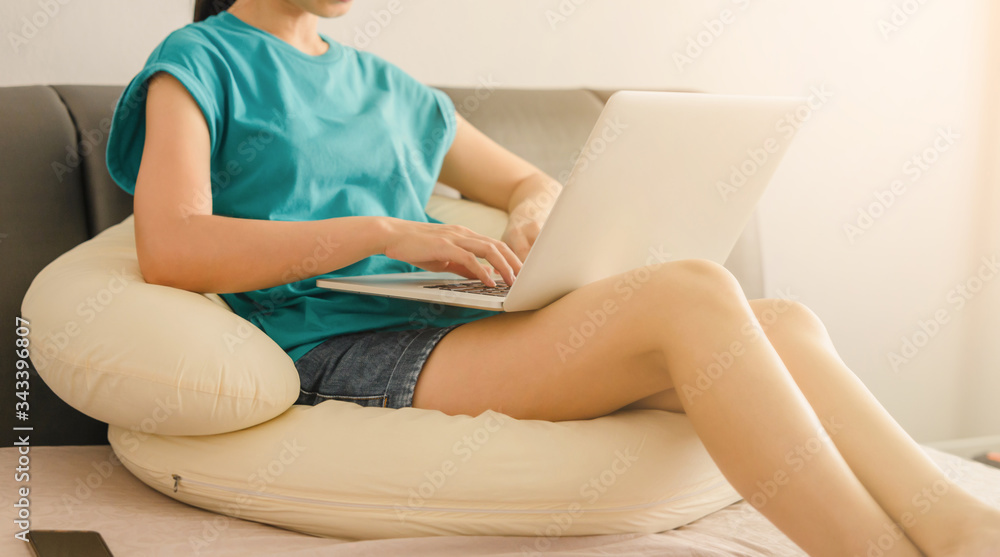 Naklejka premium Young asian woman sitting on the bed checking social apps and working on laptop computer in the bedroom while working from home. stay at home concept.