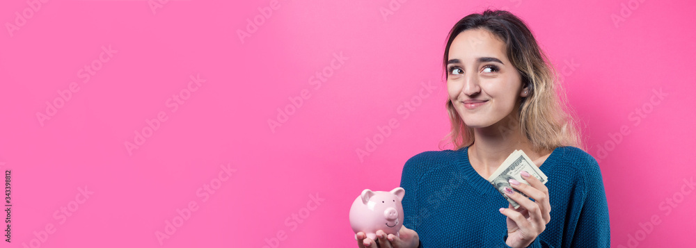 Girl puts in the piggy bank one hundred dollar bills. Young girl over pink background holding piggy bank and rejoices gesturing. The concept of reliability of cash investments and insurance.