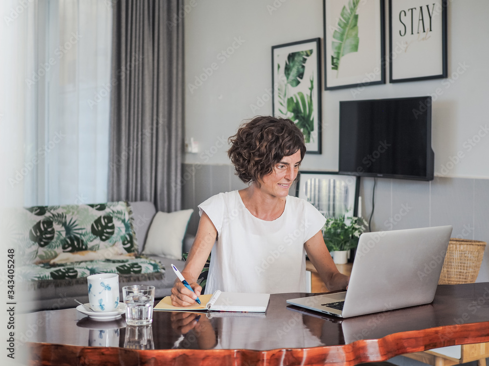 happy smiling remote online working woman with laptop, taking notes in a notebook and glass in casual outfit sitting on a work desk in her living room in her home office happy smiling remote online working woman with laptop, taking notes in a notebook and glass in casual outfit sitting on a work desk in her living room in her home office