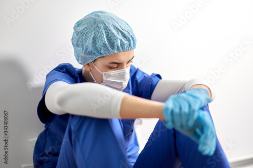 medicine, healthcare and pandemic concept - sad young female doctor or nurse wearing face protective mask for protection from virus disease sitting on floor at hospital