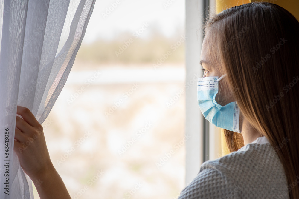 Concept of coronavirus quarantine. Child wearing medical protective face mask during flu virus, looking out of window. COVID-19 - self isolation. Teen girl forced to stay at home. Prevention epidemic.