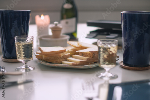 Dinner table, with slices of raclette cheese, a raclette machine, two glasses and a bottle of white wine, candle, ceramic mugs and pepper, ready for a nice traditional and typical swiss dinner.