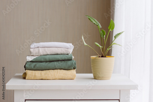 a neat stack of folded women's clothing in natural colors on a white wooden chest of drawers, a green houseplant near the window with white tulle