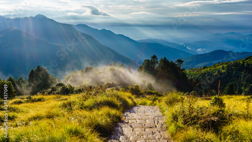 Stone paved trekking trail near the top of Poon Hill, Ghorepani in Nepal.  Fog, hills, rays of lights during sunrise in  the background..  photo