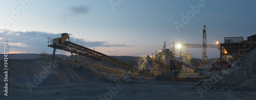 Rock stone crushing machine, conveying crushed rocks at the mining enterprise, illuminated by spotlights on a background of the dawn sky, panorama. Heavy mining equipment.