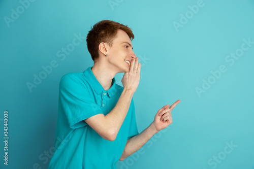 Pointing, whispering. Caucasian young man's modern portrait isolated on blue studio background, monochrome. Beautiful male model. Concept of human emotions, facial expression, sales, ad, trendy.