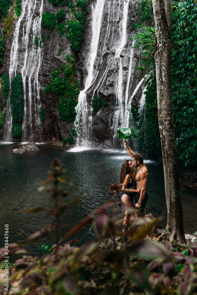Fototapeta premium Beautiful couple at a waterfall in Indonesia. A couple in love travels around the island of Bali. A man and a woman kiss at the waterfall. Travel to Asia. Honeymoon trip. Tourists at the waterfall
