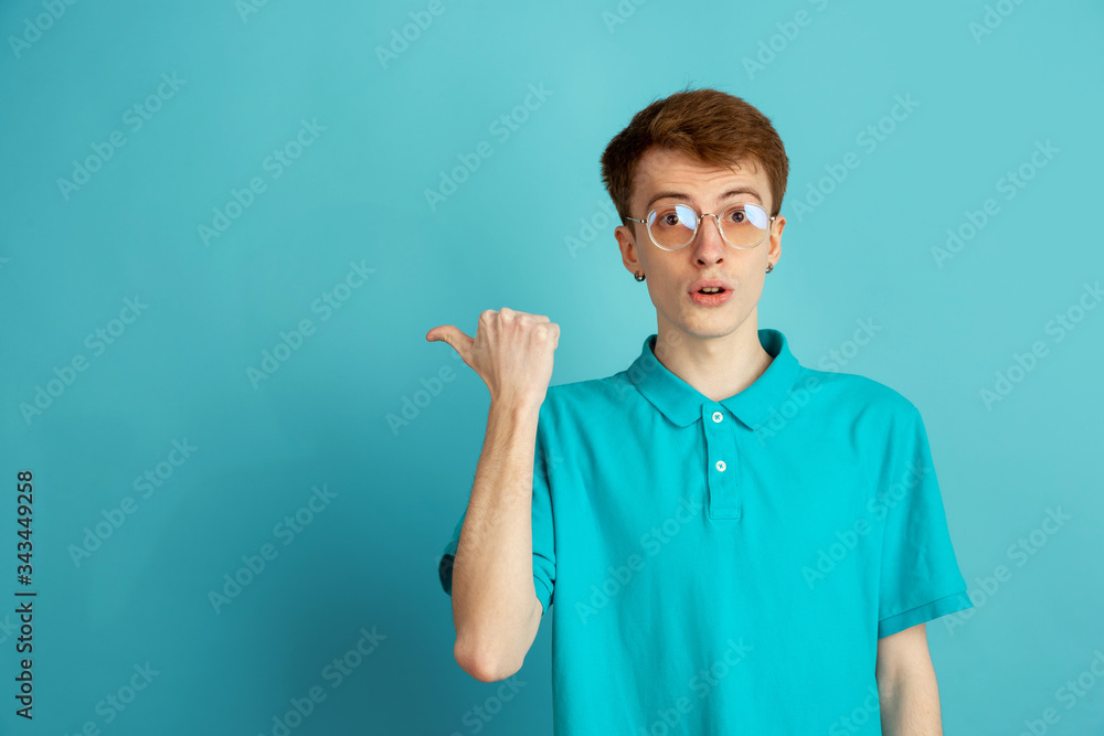 Pointing at side. Caucasian young man's modern portrait isolated on blue studio background, monochrome. Beautiful male model. Concept of human emotions, facial expression, sales, ad, trendy. Pointing at side. Caucasian young man's modern portrait isolated on blue studio background, monochrome. Beautiful male model. Concept of human emotions, facial expression, sales, ad, trendy.