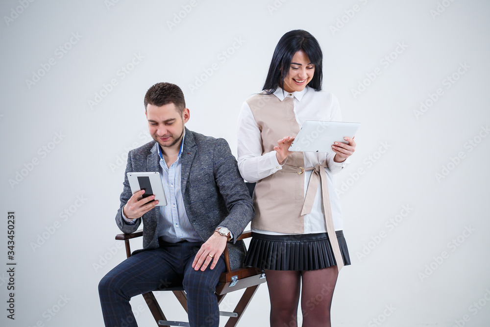 Fototapeta premium A successful man in a suit sits on a black chair and a woman stands next to look at the tablets. Future businessmen are smiling and talking about a new project.