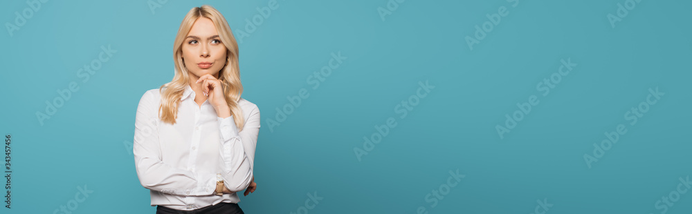 panoramic crop of dreamy businesswoman holding hand near chin while looking away on blue background panoramic crop of dreamy businesswoman holding hand near chin while looking away on blue background