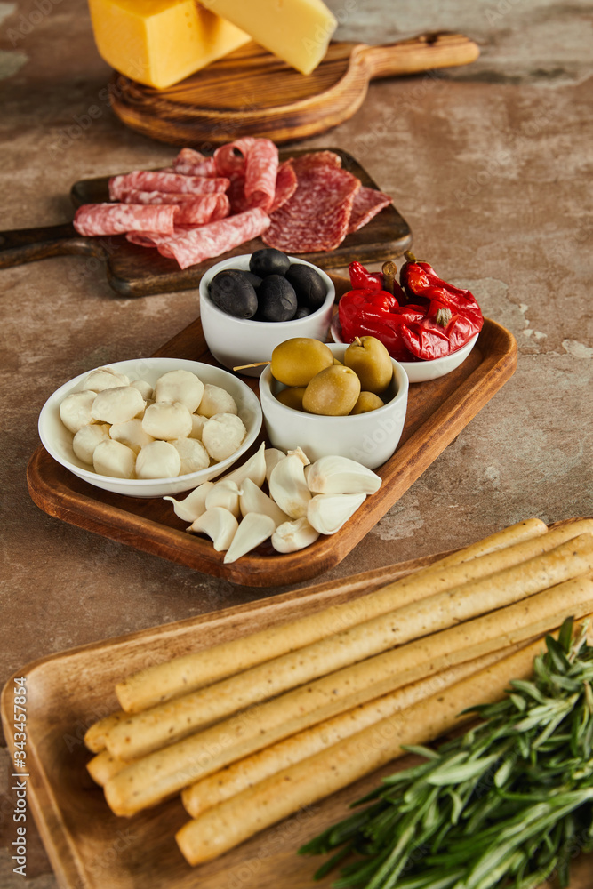 High angle view of boards with breadsticks, salami, rosemary, cheese and bowls with antipasto ingredients on brown