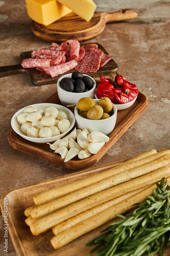 High angle view of boards with breadsticks, salami, rosemary, cheese and bowls with antipasto ingredients on brown