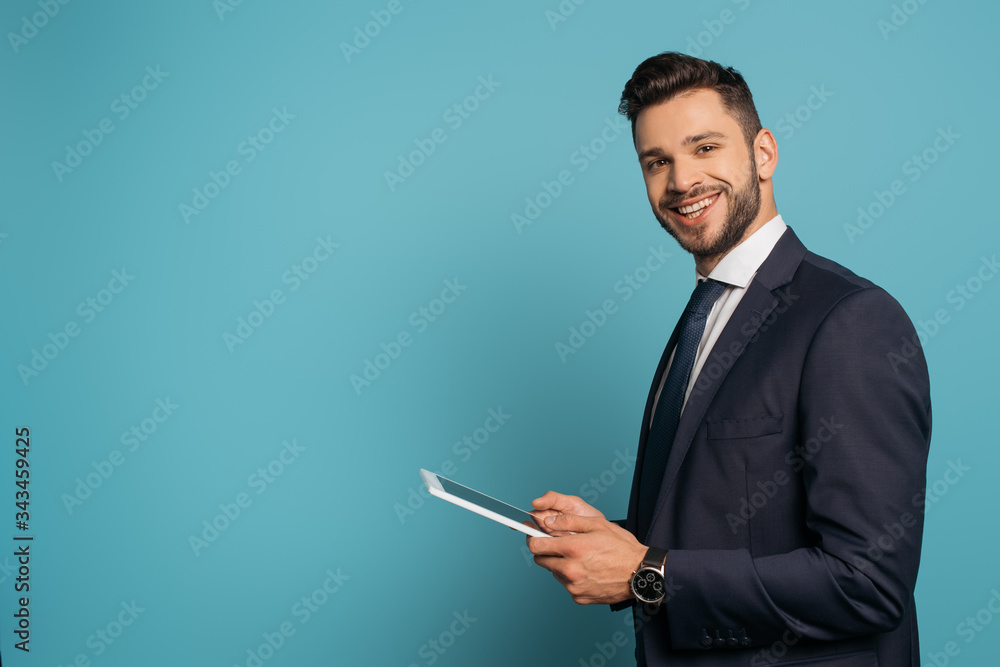handsome businessman using digital tablet while smiling at camera on blue background handsome businessman using digital tablet while smiling at camera on blue background