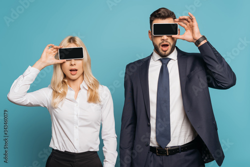 surprised businessman and businesswoman covering eyes with smartphones with blank screen on blue background