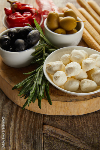 Selective focus of bowls with olives, chili peppers and mozzarella near breadsticks and greenery on board on wooden background