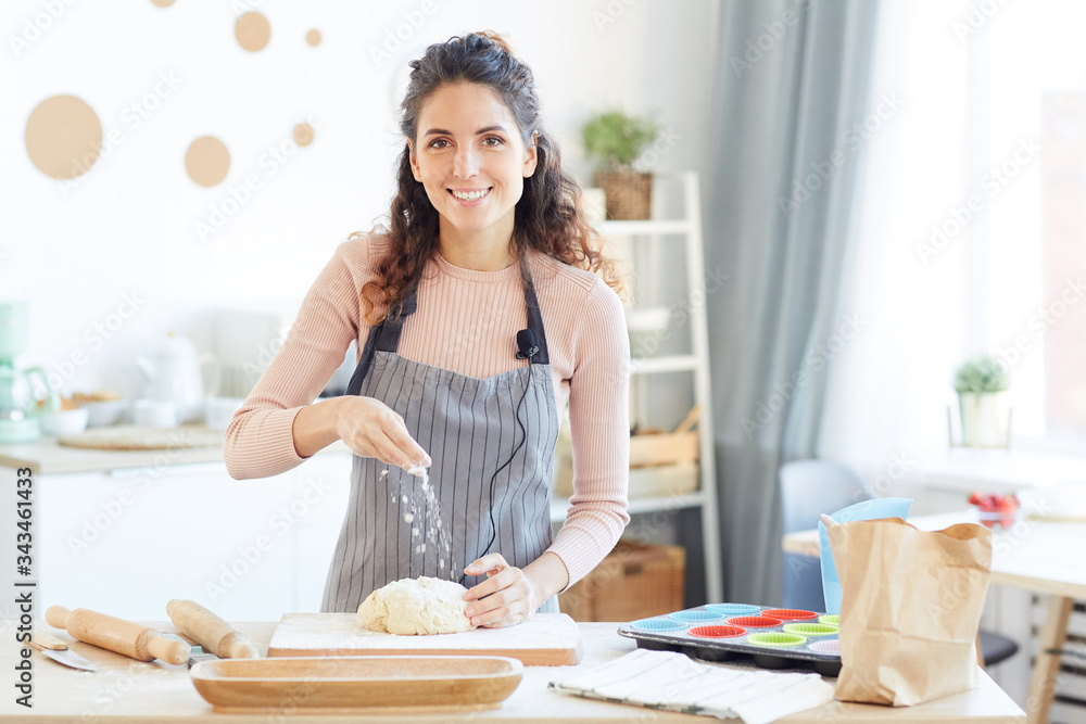 Good-looking Caucasian woman wearing apron standing at kitchen table adding flour to dough looking at camera Good-looking Caucasian woman wearing apron standing at kitchen table adding flour to dough looking at camera