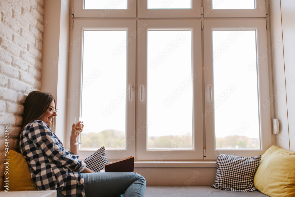 A beautiful caucasian young woman with a glass of wine by the window reading a book A beautiful caucasian young woman with a glass of wine by the window reading a book